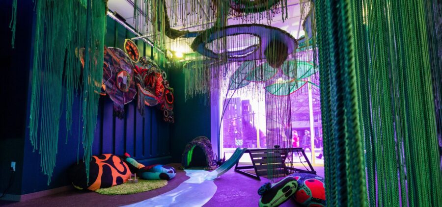 Colorful, immersive PNC PlaySpace with hanging green strands, large fabric art pieces, beanbags, a small slide, and purple lighting near a window.