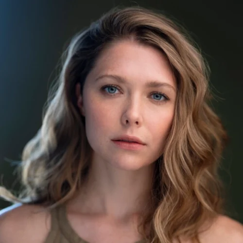 A woman with wavy blonde hair and blue eyes looks directly at the camera with a neutral expression, set against a blurred dark background.