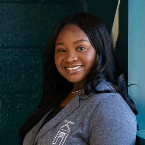 Madeleine Oldham, a woman with long black hair, wearing a gray blazer and pearl necklace, smiles while standing against a dark green wall.