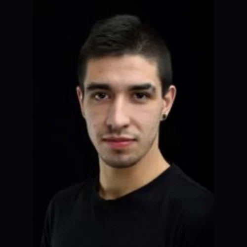 A young man with short dark hair, light skin, and a trimmed beard, wearing a black shirt and black earring, poses against a black background.