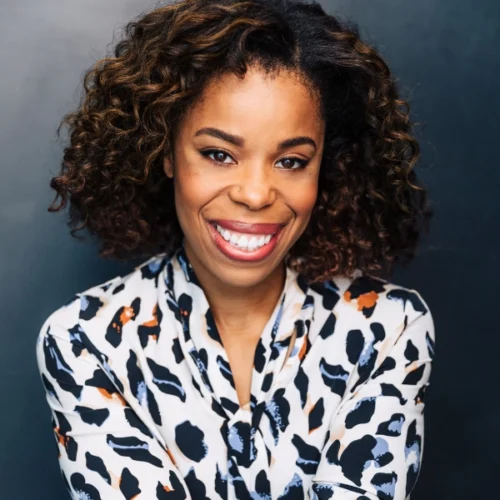 A woman with curly hair smiles at the camera, wearing a patterned blouse against a plain dark background.