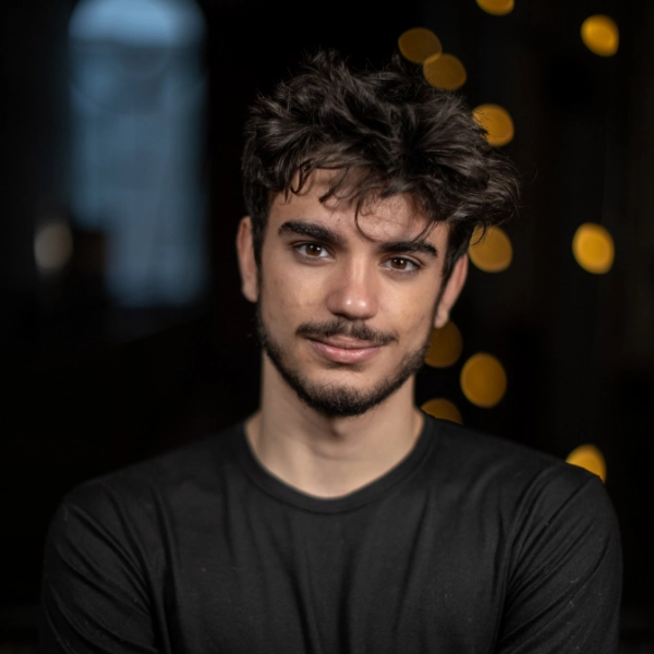 A young man with dark hair and facial hair stands indoors, wearing a black shirt, with blurred yellow lights in the background.