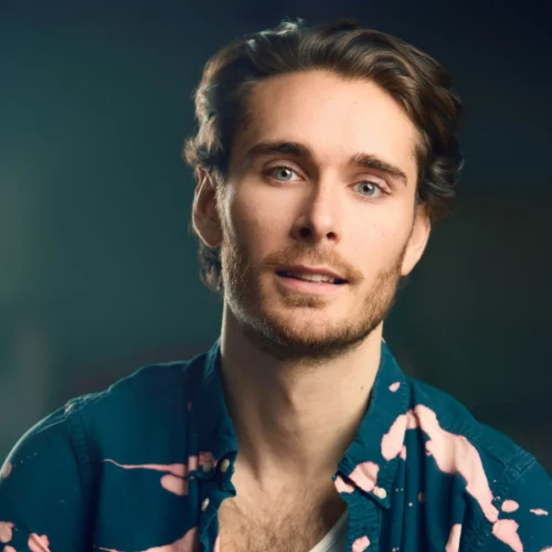 A young man with light skin, brown hair, and a short beard wearing a dark blue shirt with pink patterns, looks slightly past the camera against a blurred background.