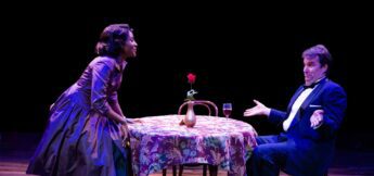 Two actors perform on stage at a table with a floral cloth, a vase with a rose, and a wine glass. The woman stands, leaning slightly forward, while the man sits with an expressive gesture.