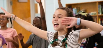 A group of middle school campers stand indoors, raising their arms and smiling enthusiastically, with bookshelves and a door visible in the background at Alliance Theatre advanced musical theater camp.