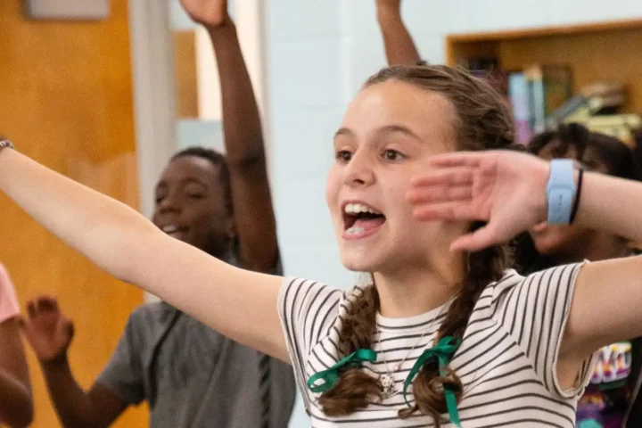 A group of middle school campers stand indoors, raising their arms and smiling enthusiastically, with bookshelves and a door visible in the background at Alliance Theatre advanced musical theater camp.