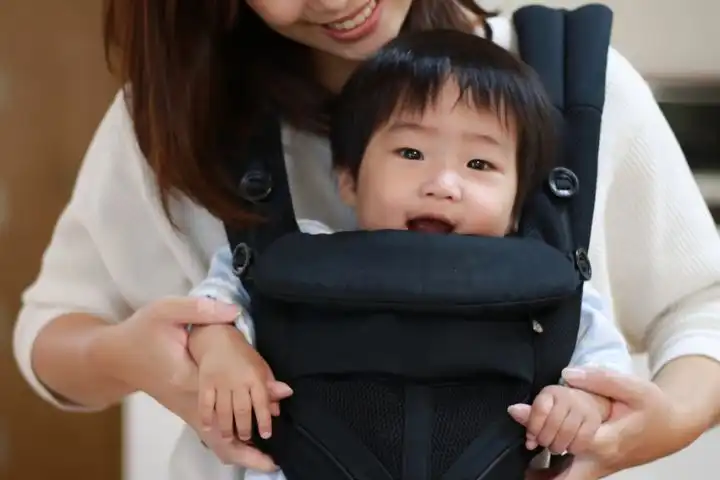A smiling baby sits in a front-facing black carrier while an adult, partially visible, supports them from behind.