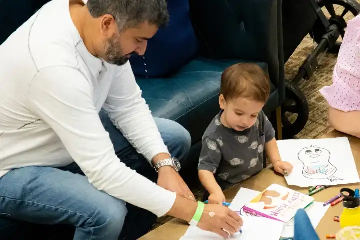 An adult and a young child sit at a table drawing with markers and crayons on white paper, with finished drawings visible on the table.