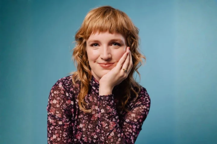 Announcing the Cast and Creative Team of Fires Ohio: A person with wavy blond hair and bangs, wearing a floral top, smiles with their chin resting on their hand in front of a plain blue background.