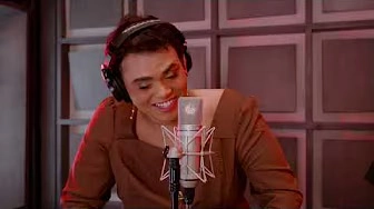 Person wearing headphones and a brown top, speaking or singing into a microphone—like father, like child—in a recording studio with gray soundproof panels.