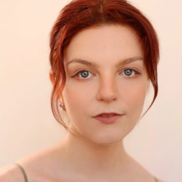 Caroline Pernick, a young woman with fair skin and red hair, looks directly at the camera with a neutral expression, set against a plain light background.