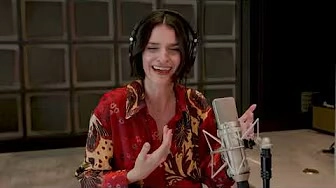 A woman wearing headphones and a red patterned shirt speaks and gestures while sitting in front of a microphone in a recording studio, perhaps sharing stories inspired by Like Father.