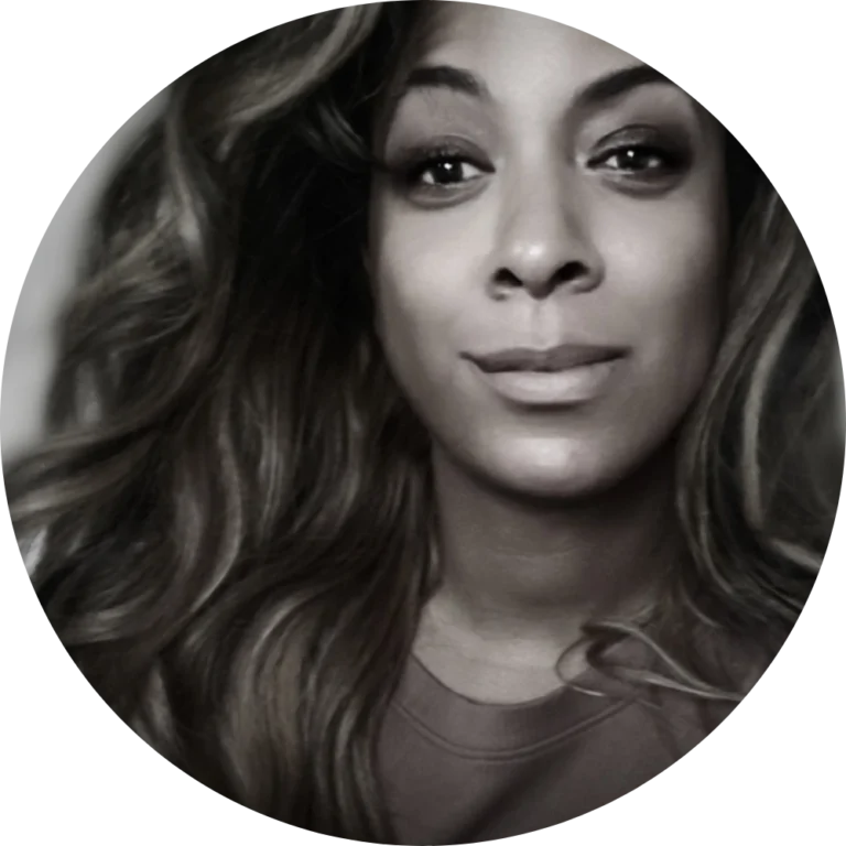 A person with long, wavy hair and a neutral expression looks at the camera, evoking a sense of quiet Community Conversation. The image is in black and white with a circular crop.