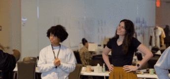 Two people stand in a rehearsal room; one looks forward with hands clasped, as if awaiting intro directing theater notes, while the other stands with hands on hips, smiling. Desks and papers are visible in the background.