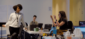 Three women engage in a lively discussion in a rehearsal room, laptops open and papers scattered, as they rehearse a show.