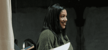 A woman with long dark hair wearing a green sweater sits indoors, smiling and looking to her left, appearing relaxed.