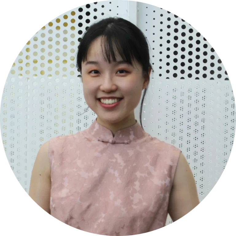 A woman in a sleeveless, light pink patterned top stands in front of a white perforated metal background, smiling at the camera during Alliance/Kendeda Week: The Words of Ants.