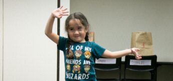 Young girl with braided hair stands indoors in Broadway Dance class, smiling with one arm raised. She wears a blue shirt that says "All of my friends are wizards." Paper bags and chairs are in the background.