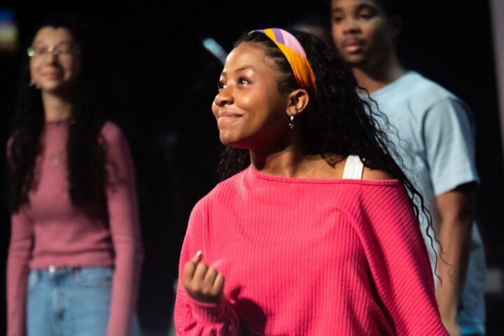 A young woman in a pink top and headband stands in focus, smiling, with two other people blurred in the background on a stage or indoors.