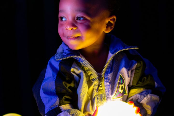 A young child in a blue jacket holds a glowing orb, illuminated by its light in a dark setting.