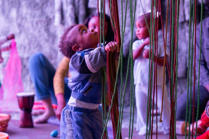 A young child stands and holds colorful hanging ropes, while other children and an adult sit or stand in the background in a play area.