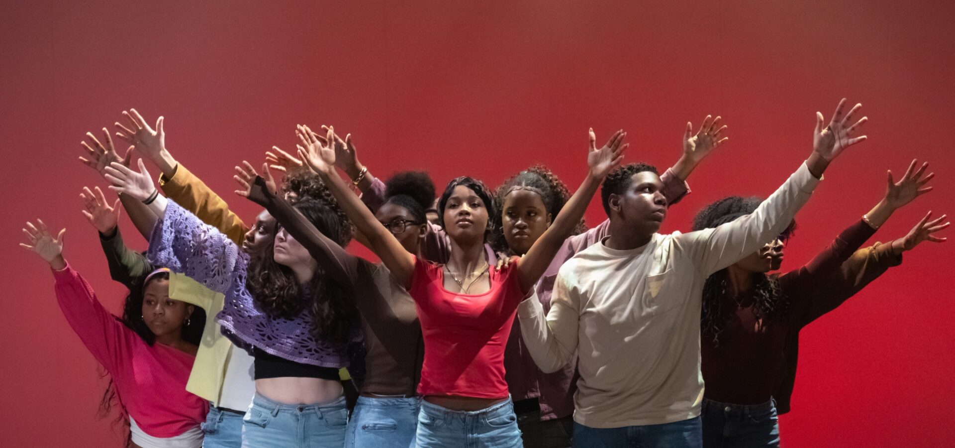 A group of people in casual clothes stand closely together against a red background, raising their arms in various directions.
