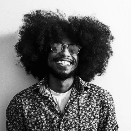 A smiling man with a large afro hairstyle, glasses, and a patterned shirt stands against a plain white background.