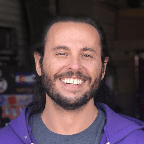 A man with long dark hair and a beard smiles at the camera, wearing a purple hoodie and a grey t-shirt, with a blurred indoor background.