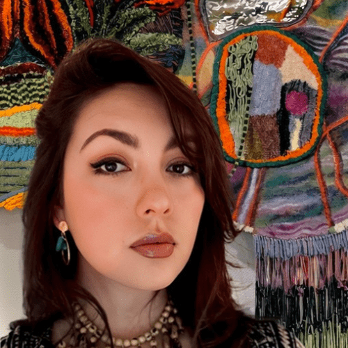 A woman with long brown hair and jewelry stands in front of a colorful, textured textile artwork featuring abstract patterns.