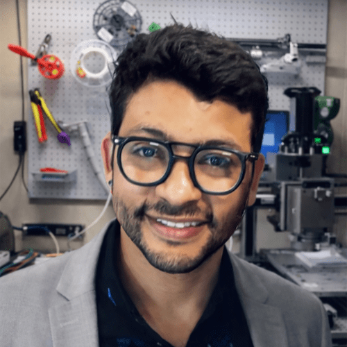 A man wearing glasses and a gray blazer smiles at the camera in a workshop with tools and equipment in the background.