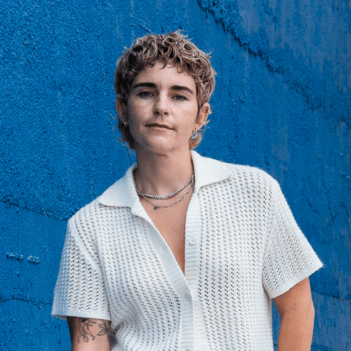 Person with short curly hair and tattoos wearing a white textured shirt stands in front of a bright blue textured wall, looking at the camera.