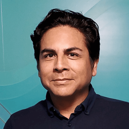 A man with dark hair wearing a navy collared shirt stands in front of a teal background with a subtle geometric pattern.