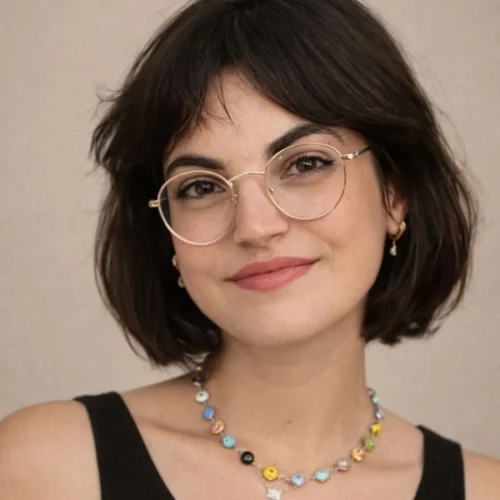 Woman with short dark hair, round glasses, and a sleeveless black top, wearing a colorful beaded necklace, smiling at the camera against a neutral background.