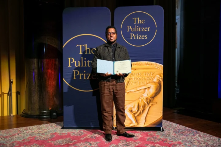 A person stands with a sense of purpose, holding a certificate in front of a backdrop with "The Pulitzer Prizes" logo at an award event.