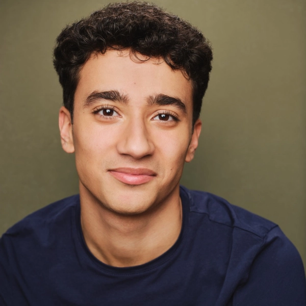 Coty Perno, a young man with short curly hair, wears a navy blue shirt and smiles slightly while facing the camera, set against a plain olive green background.
