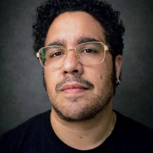 A person with curly dark hair, glasses, a mustache, and beard stubble looks directly at the camera against a dark background.