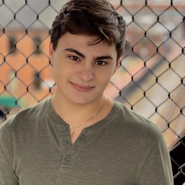 A young person with short brown hair, wearing a green henley shirt and gold necklace, stands in front of a chain-link fence with blurred buildings in the background.