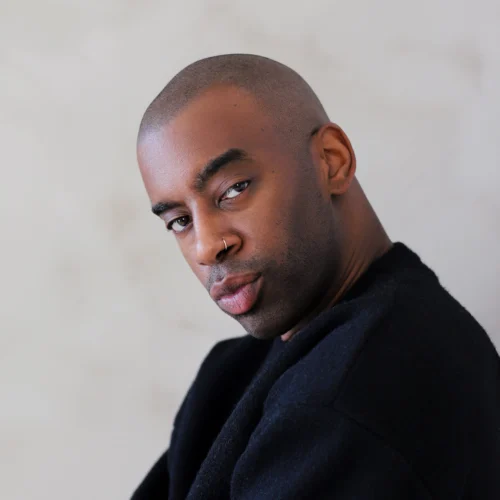 Dominique Kelley, a man with a shaved head and trimmed beard, wears a black sweater as he sits against a light-colored background, looking at the camera.