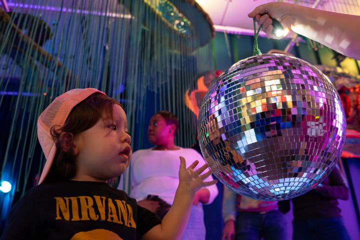 A young child wearing a Nirvana shirt and cap reaches toward a large disco ball, with two adults standing in the background under hanging decorations.