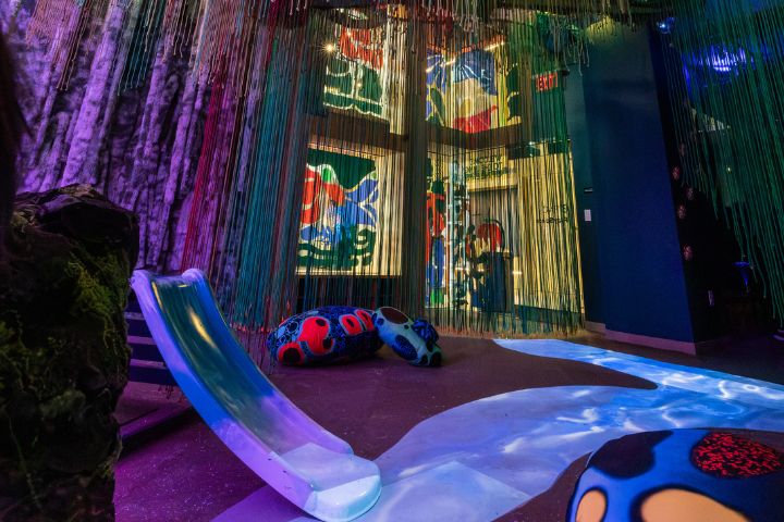 Colorful indoor play area with a slide, abstract wall art, hanging strings, and large, patterned soft play structures on the floor.