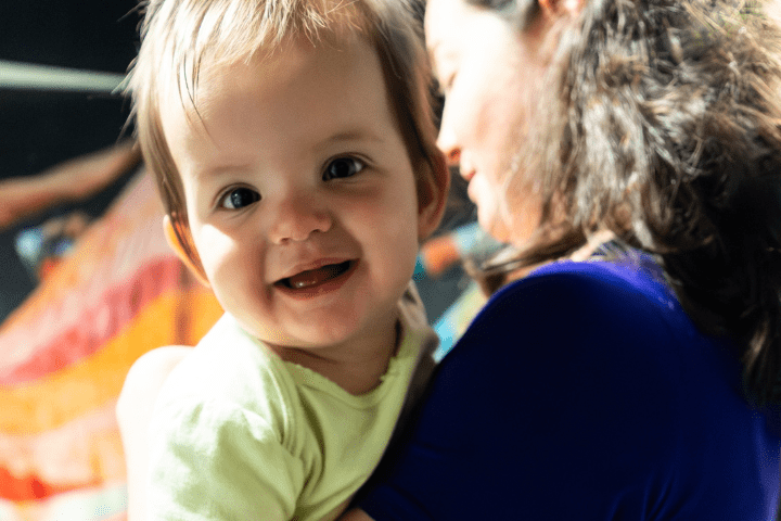 A baby smiles at the camera while being held by a woman with long brown hair, shown from the side, in a brightly lit setting.