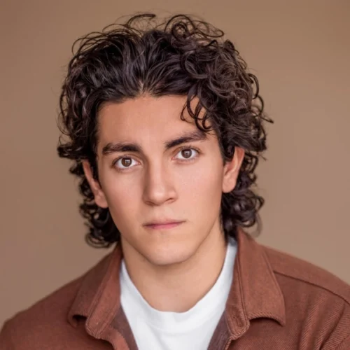 A young man with curly brown hair and brown eyes looks seriously at the camera, wearing a white shirt and a brown jacket against a plain beige background.