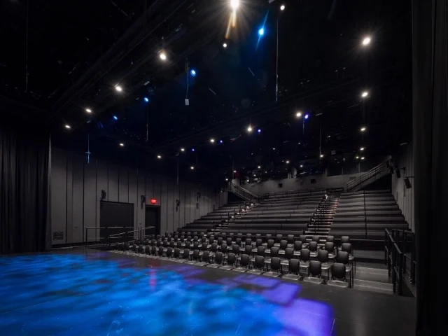 An empty black box theater with rows of black seats facing the Goizueta Stage, illuminated by blue and white stage lights from above.