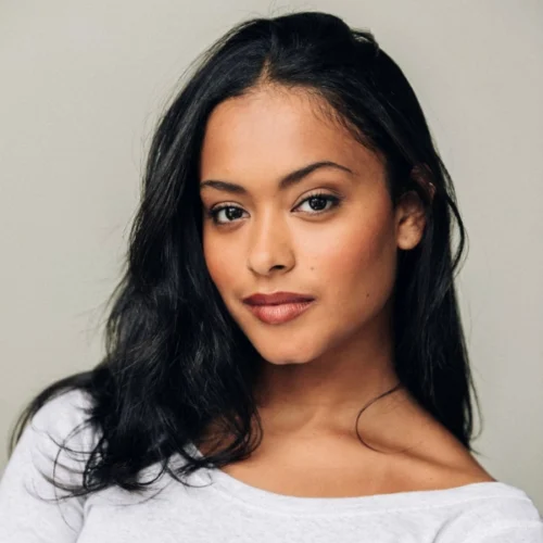A woman with long black hair and medium skin tone looks at the camera, wearing a light grey top against a plain background.