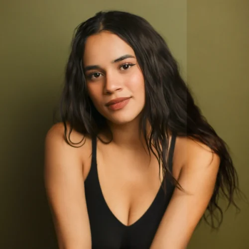 Woman with long wavy dark hair wearing a black sleeveless top poses in front of an olive green background, looking at the camera with a neutral expression.