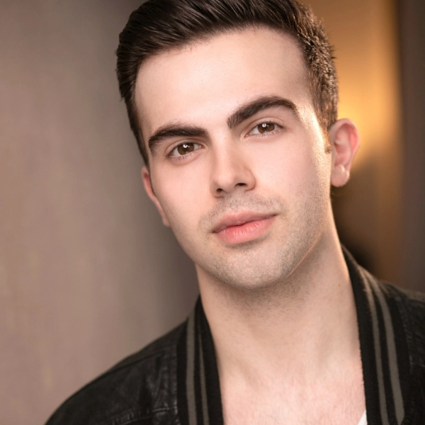 A young man with short dark hair and a trimmed beard looks at the camera, wearing a black jacket over a white shirt, with a neutral background.