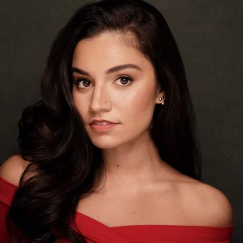 A woman with long dark hair, wearing an off-the-shoulder red top, looks directly at the camera against a dark background.
