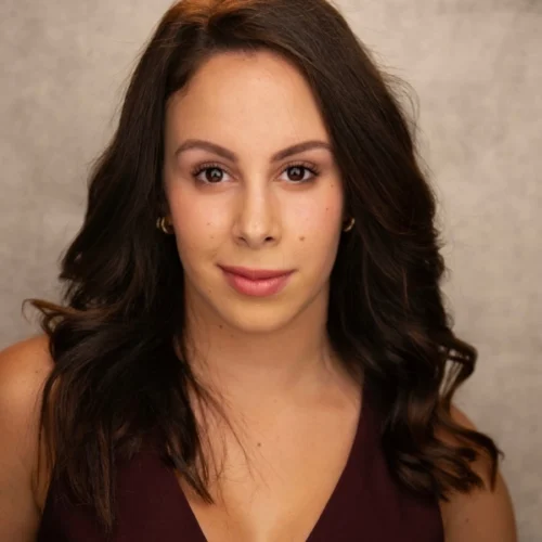 A woman with long brown hair and a maroon top looks directly at the camera against a neutral gray background.