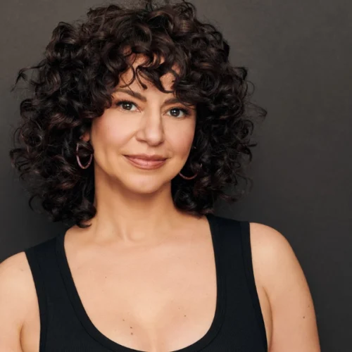 Woman with short, curly dark hair wearing a sleeveless black top, smiling slightly, standing against a plain dark background.