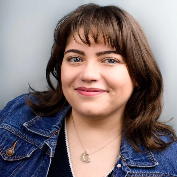 A woman with shoulder-length brown hair and bangs smiles at the camera. She is wearing a denim jacket and a necklace with a round pendant. The background is plain and light-colored.
